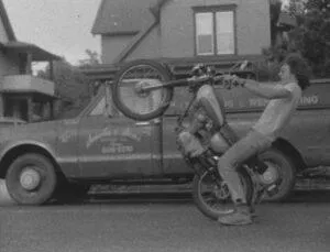 Mike Smola doing a wheelie on a motorcycle with the roofing truck behind him